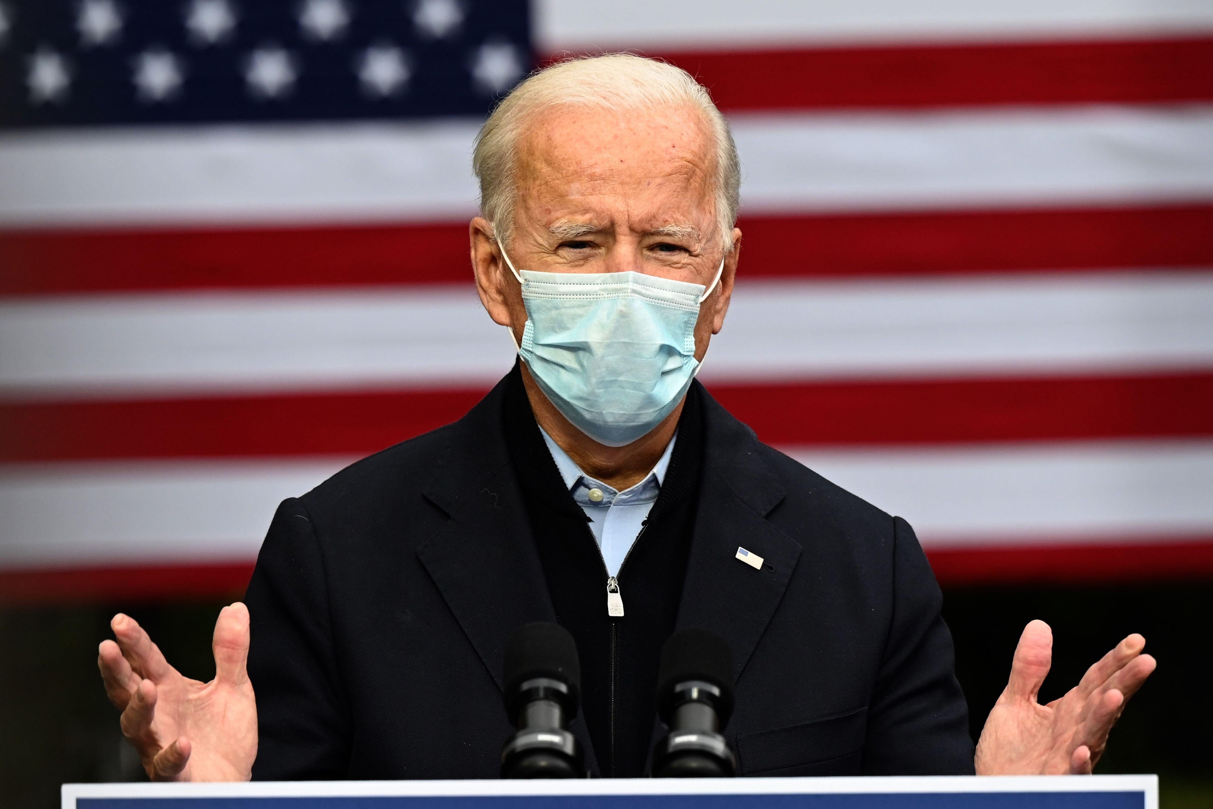 Joe Biden stands in front of a flag, wearing a surgical mask and speaking from a lectern.