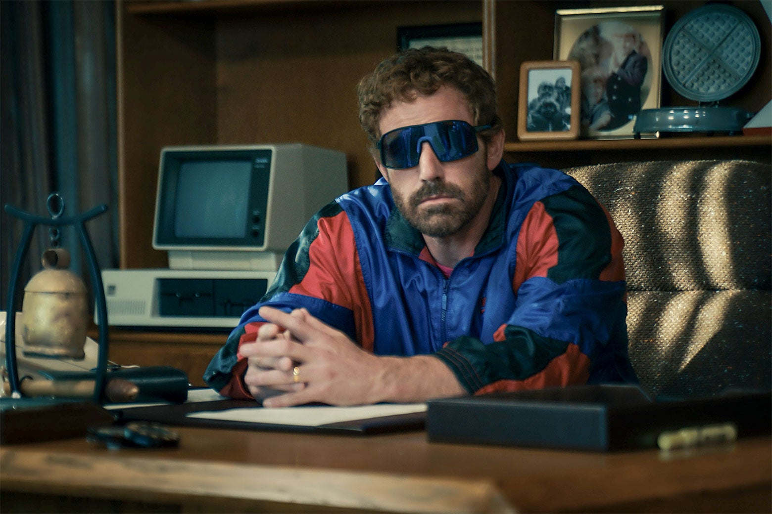 The actor sits behind an impressive desk, wearing a red, black, and blue tracksuit, curly hair, and some very goofy and extremely 1980s sunglasses.