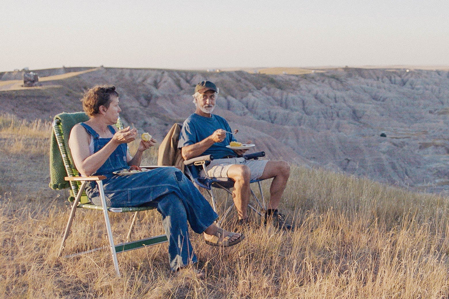 A still from Nomadland: Frances McDormand as Fern and David Strathairn as Dave sit on lawn chairs in front of a scenic backdrop.