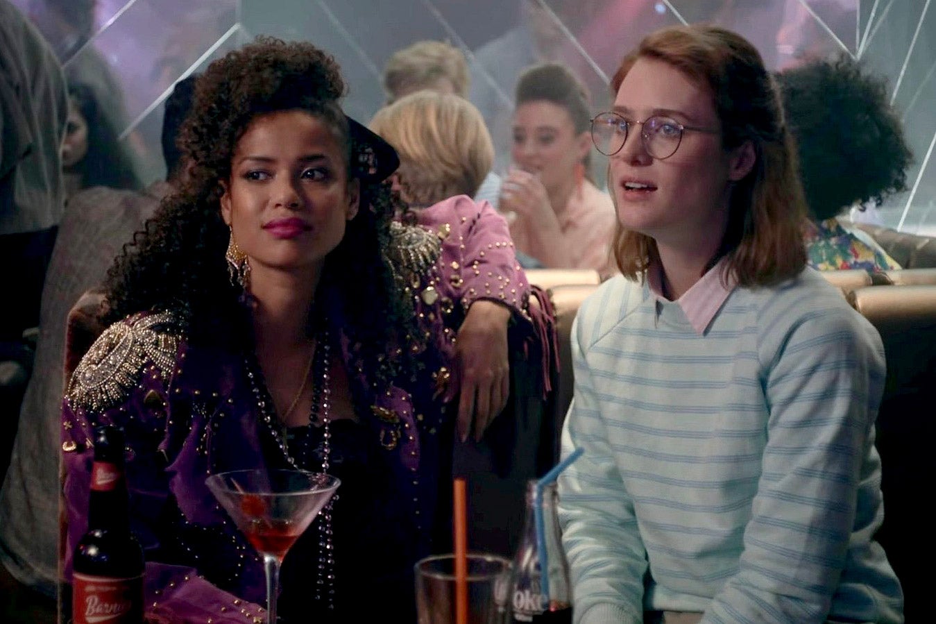 A black woman in glam ’80s gear and a white woman in preppy ’80s gear sit in a bar and look ahead in amusement.