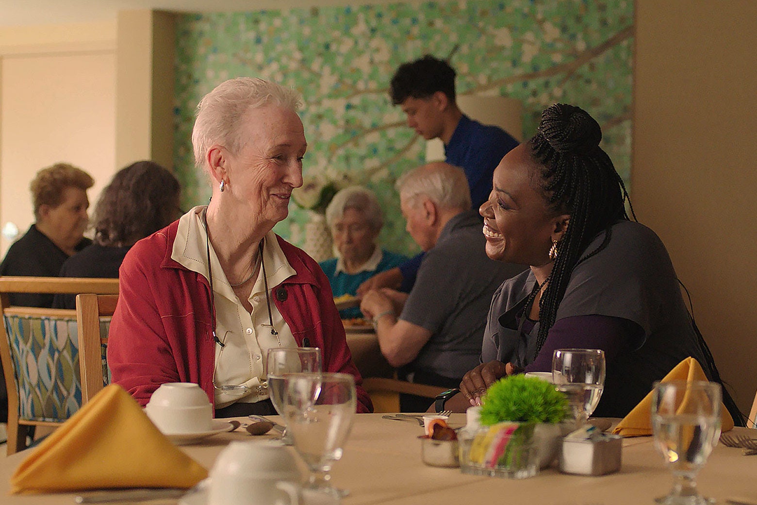 An older white woman sitting at a table smiling and chatting with a younger Black woman dressed in medical scrubs.
