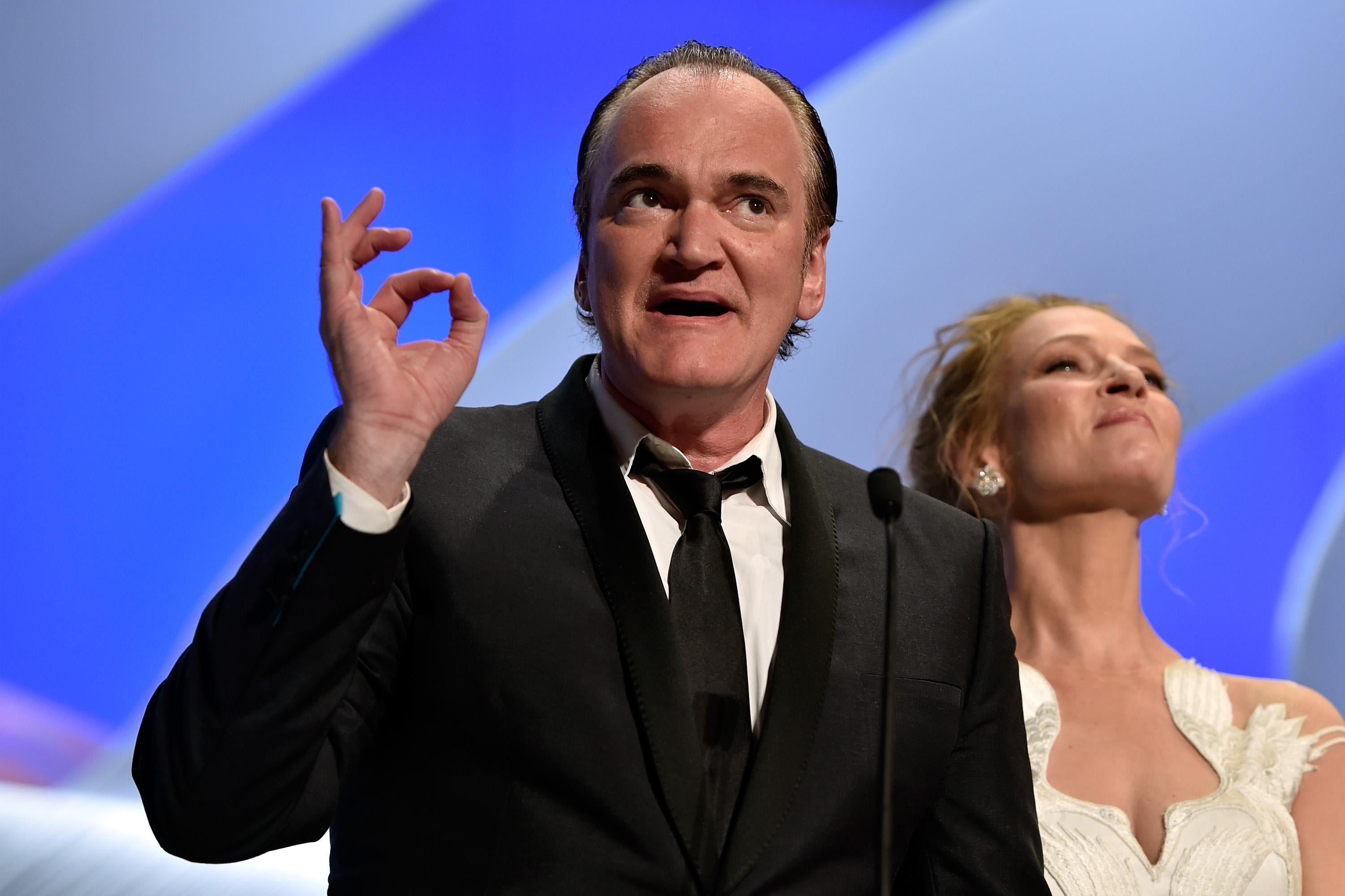 Quentin Tarantino holds his thumb and index finger together as Uma Thurman stands behind him