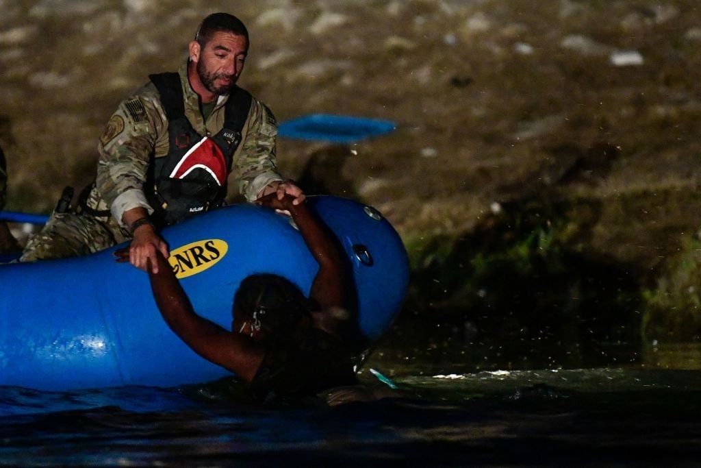 A man in a camouflage uniform in a blue inflatable boat holds the arms of a woman who is submerged up to her chest in water at night.