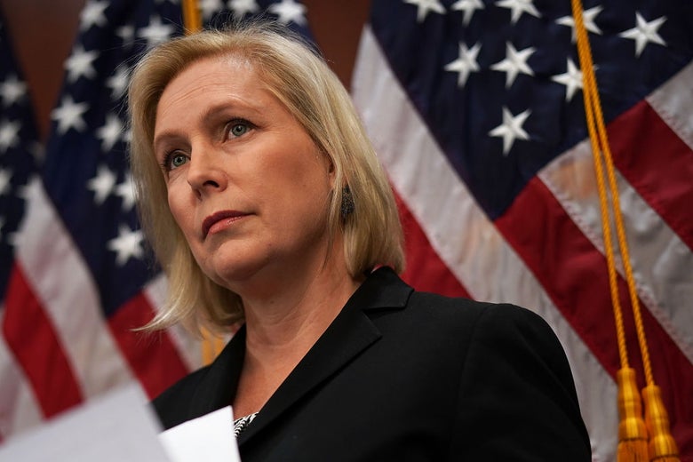 Gillibrand, holding papers and wearing a black suit, stands in front of flags.