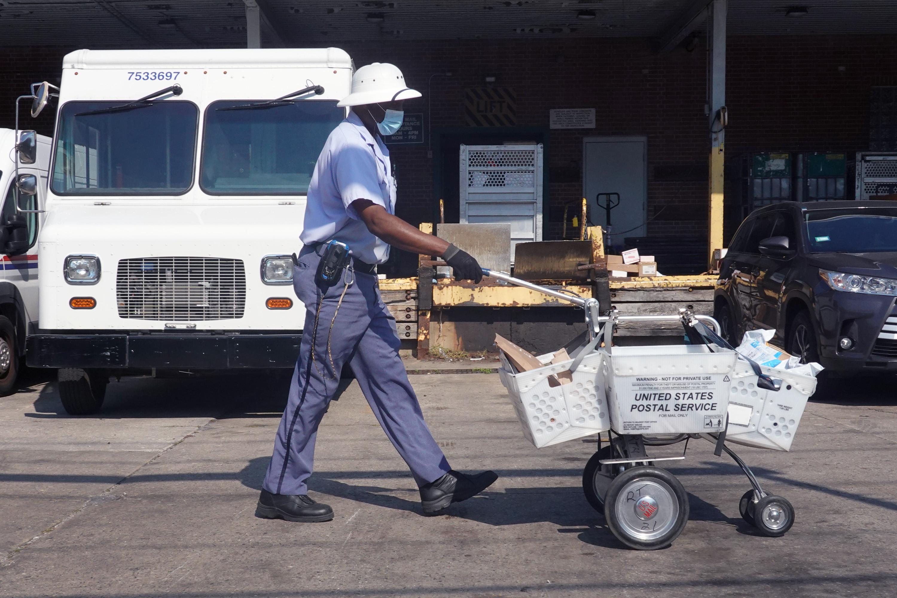 A postal worker in uniform pushes a cart full of mail past a loading dock area.