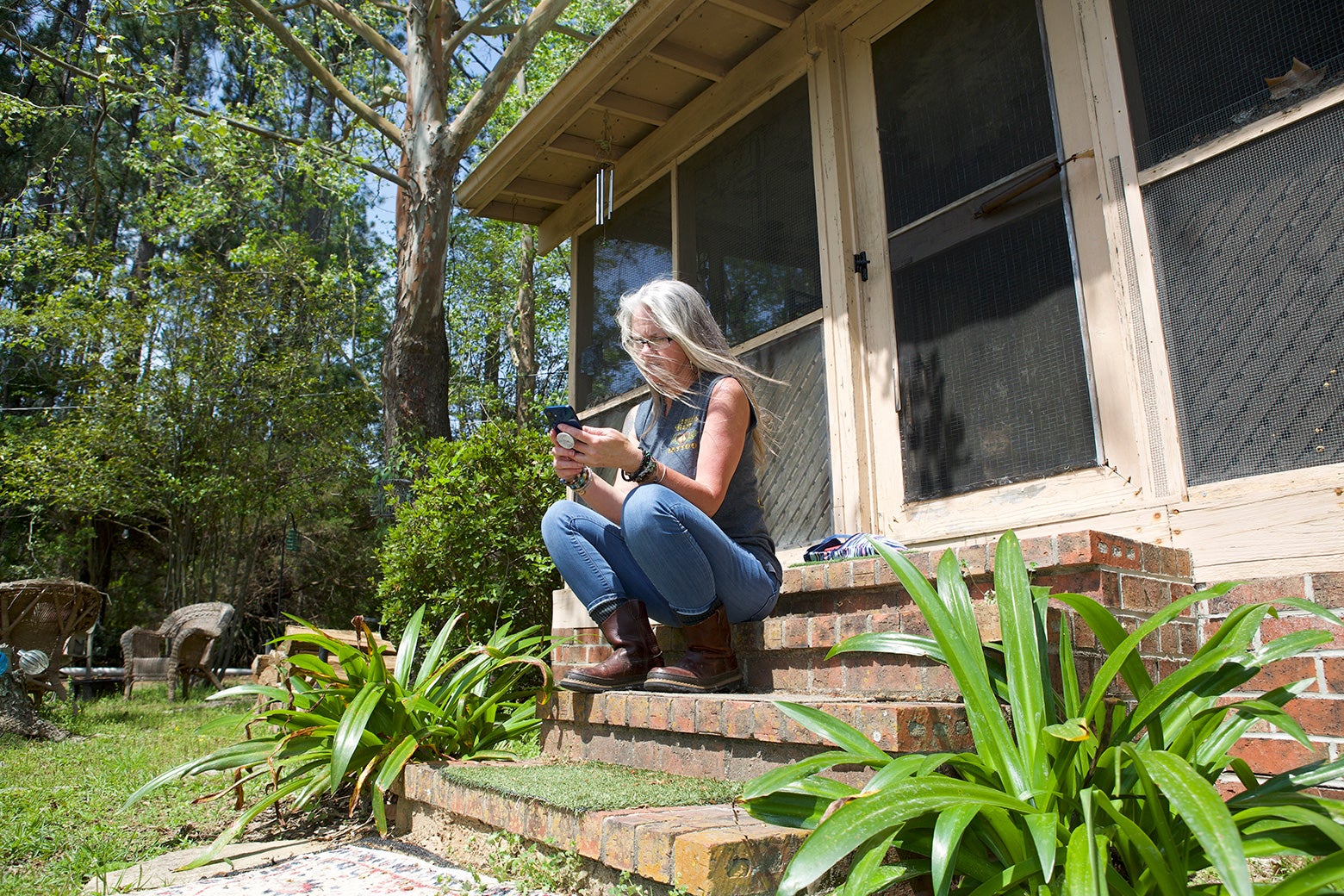 A woman with long gray hair sits on brick steps outside her house and looks down at her phone.