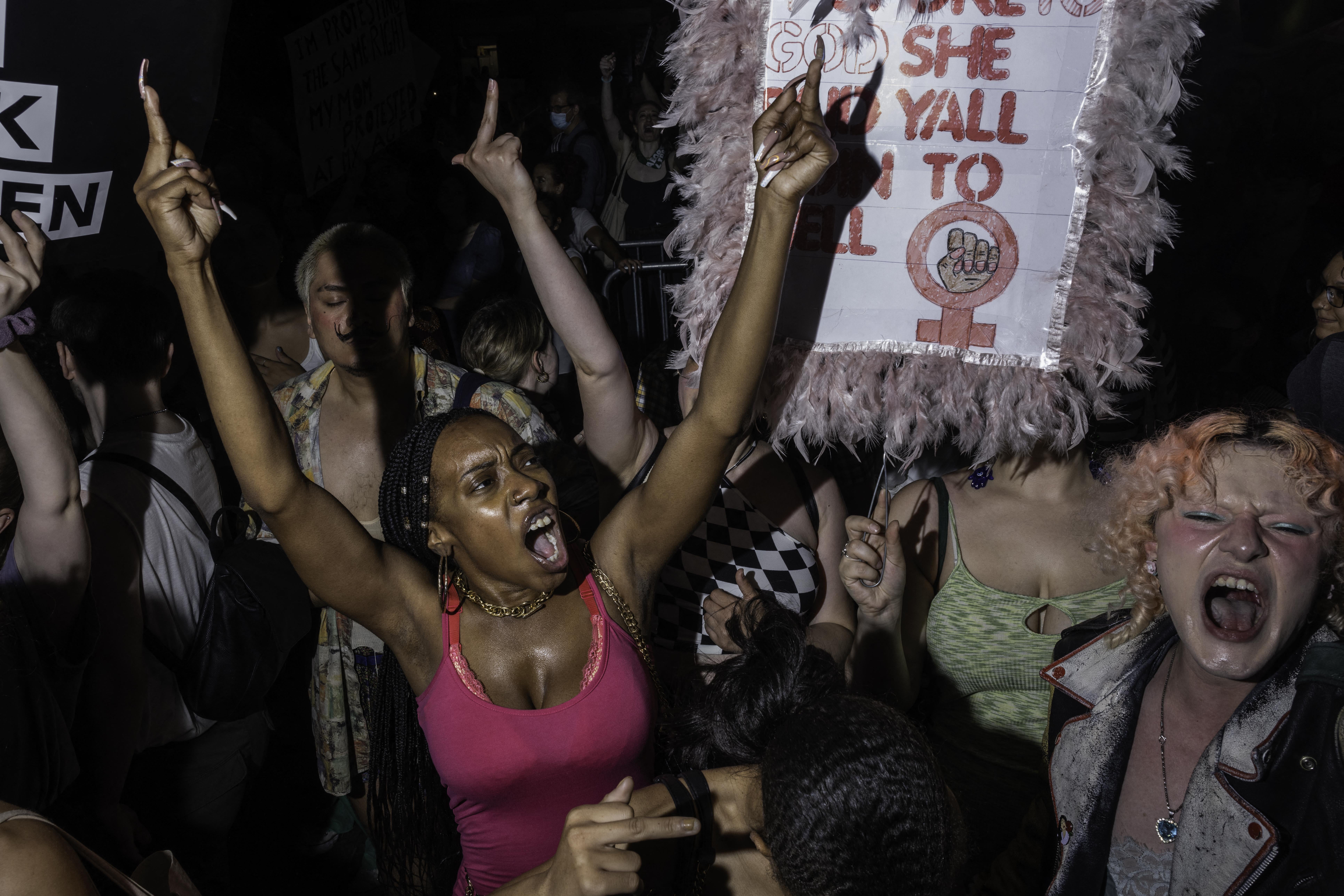 In a crowd, one woman yells while lifting her hands high; a woman next to her holds a sign.