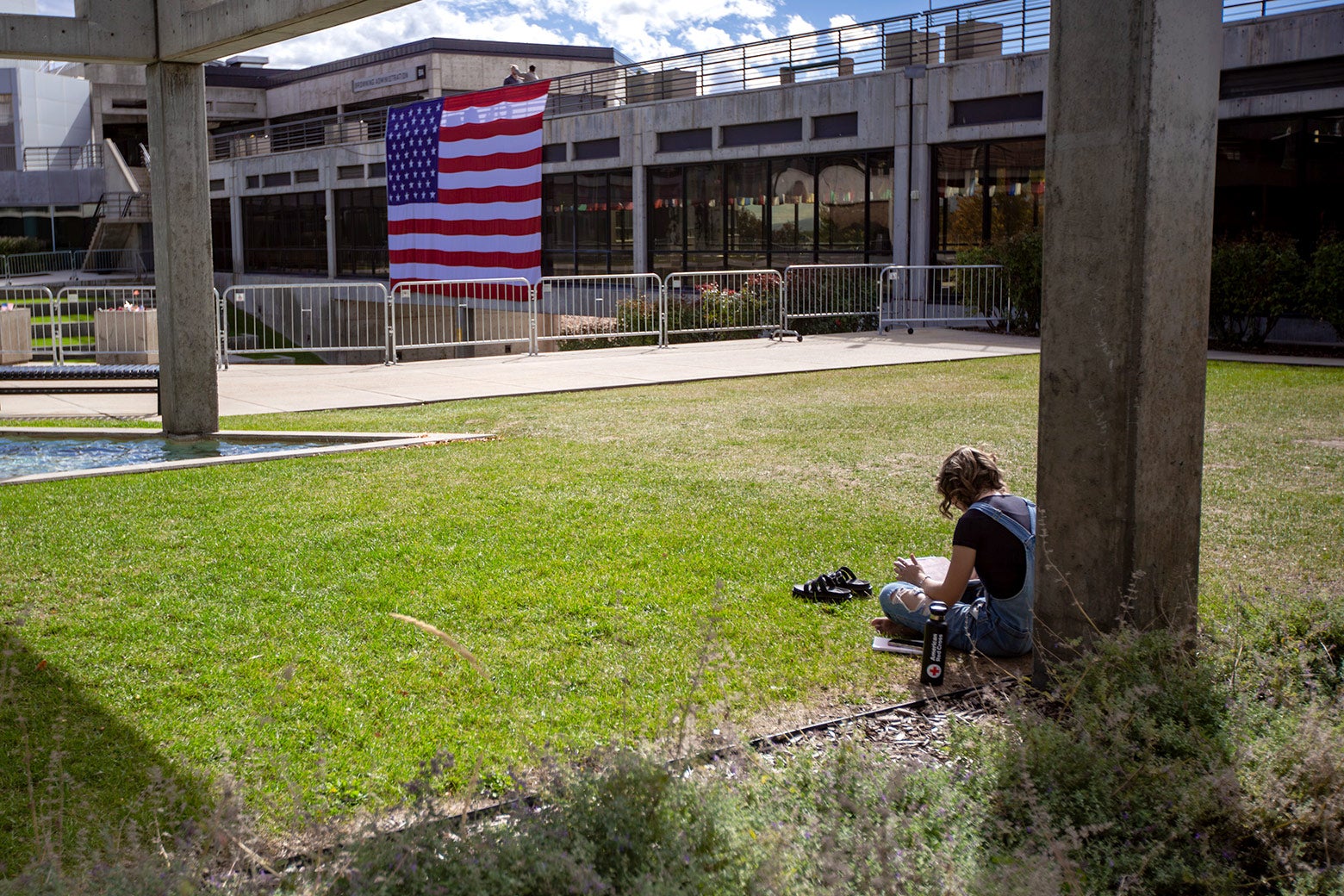 A student sits on the grass near fenced-off amphitheater with a huge American flag draped over it.