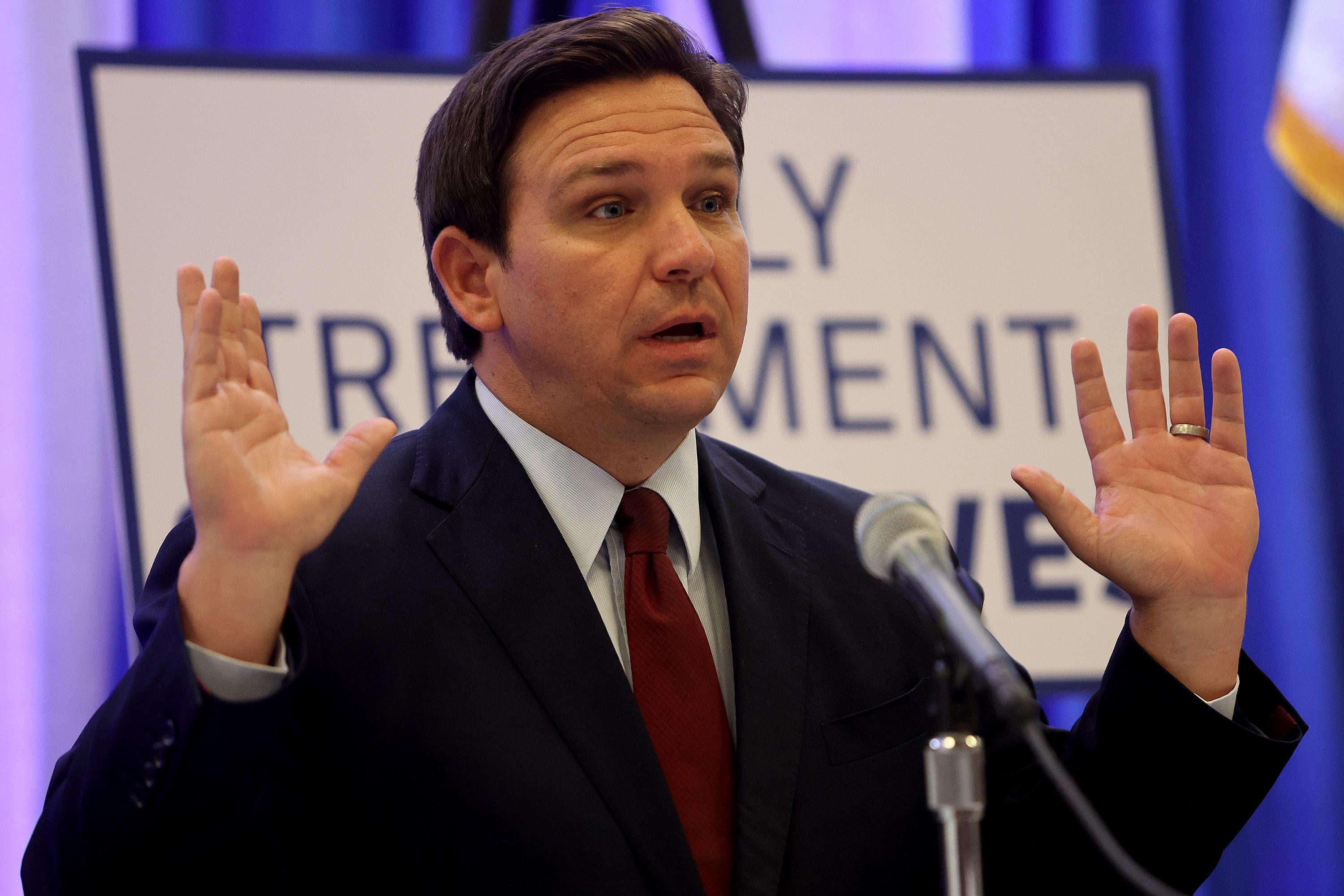 Ron DeSantis raises his hands while speaking in a mic in front of a sign.