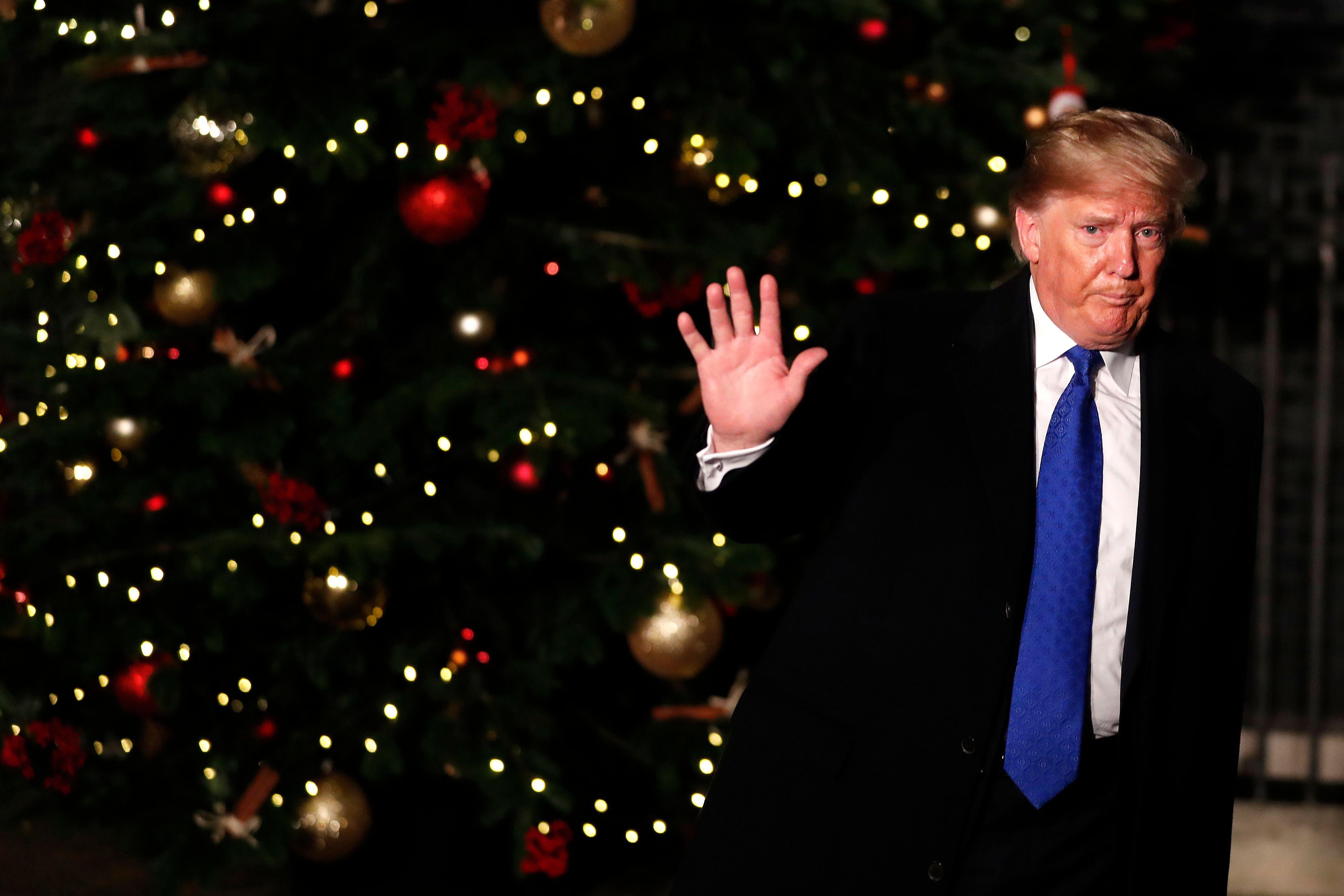 Trump is standing in front of a Christmas tree, waving goodbye, with a neutral or negative facial expression