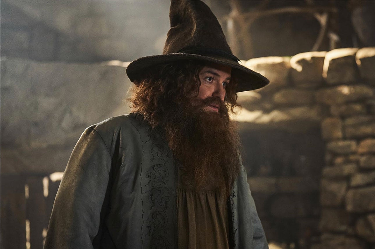 A picture of Tom Bombadil from the show; he has a (not bright) blue shirt and a long beard and a wizard's hat and is standing in front of a large stone fireplace.