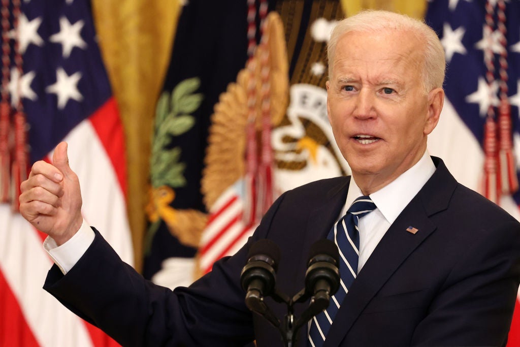 Biden, seen at a lectern against a backdrop of American and presidential-seal flags, gestures with his right hand and thumb.