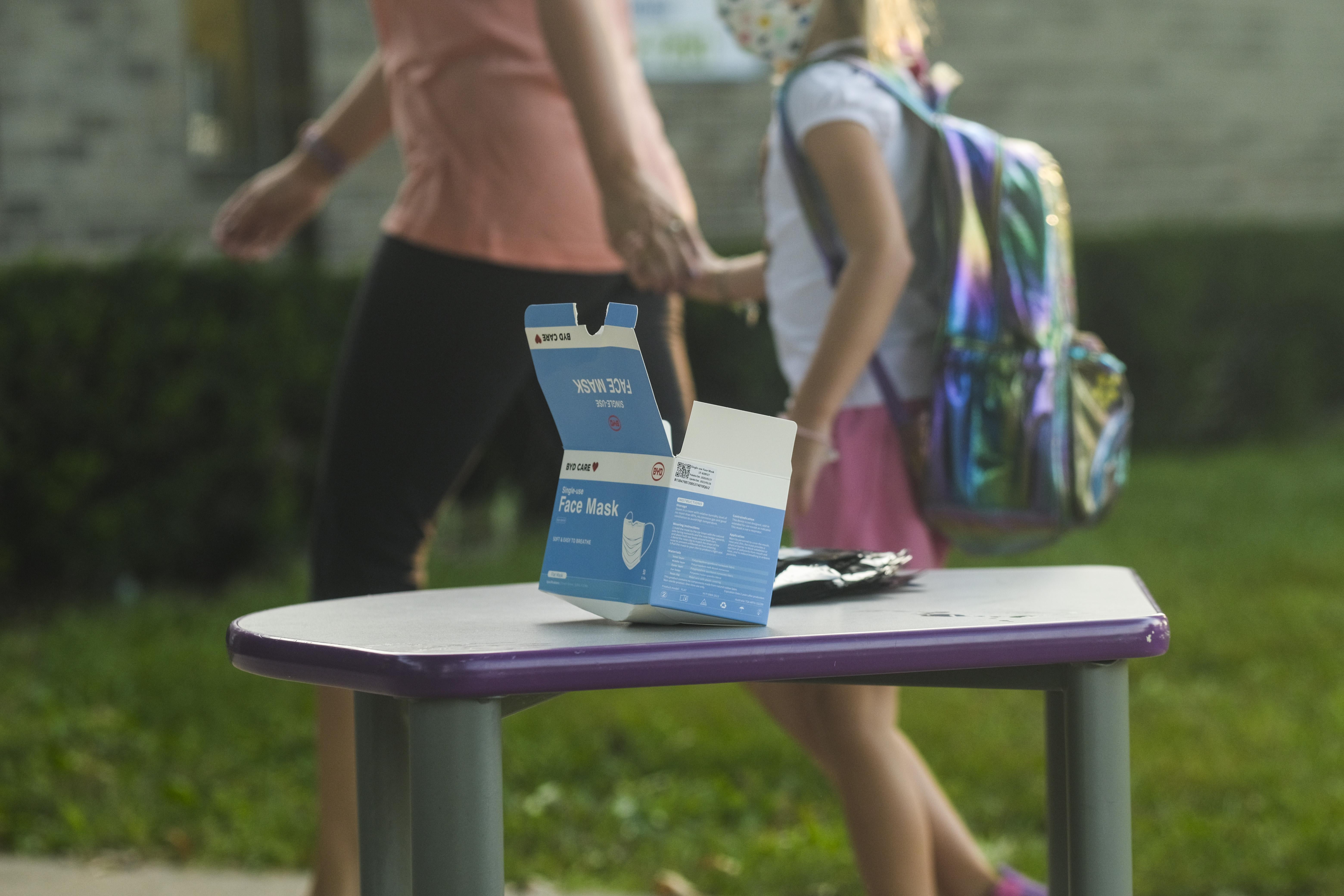 A box of face masks sits on a table outside of of a school. In the background, a parent holds their child's hand as they walk toward school. The child is wearing a face mask and backpack.