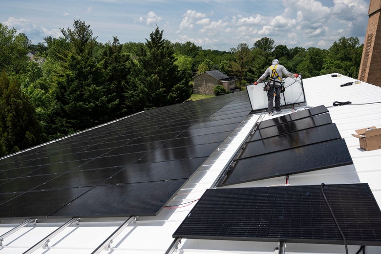 A man installs solar panels on the roof of a building surrounded by trees.