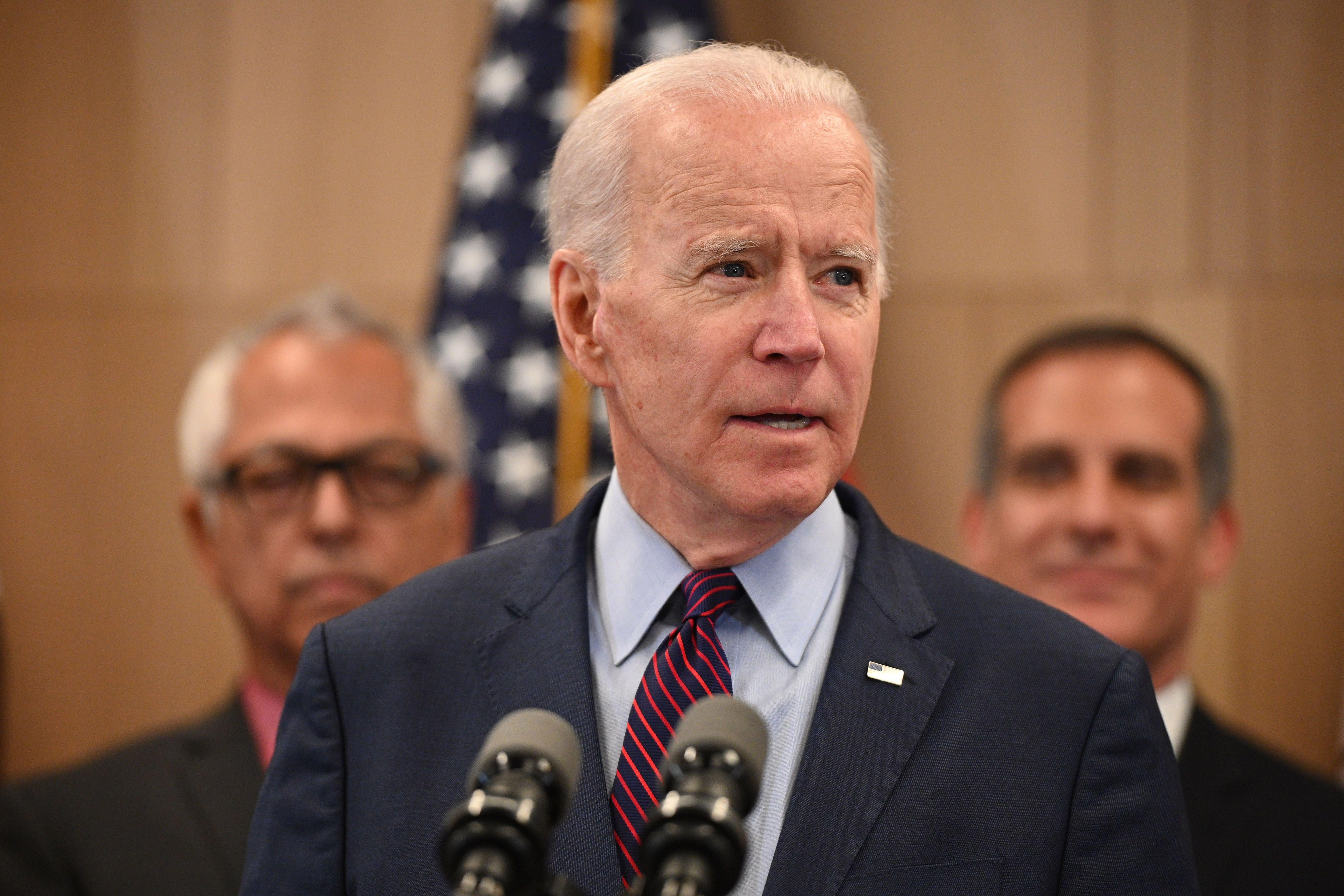 Joe Biden stands in front of two microphones, flanked by men on both sides of him.