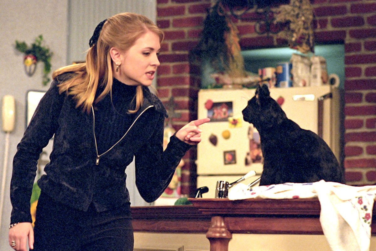 Melissa Joan Hart, dressed in black pants and a black jacket overtop a black turtleneck sweater points her finger in a chastising manner at a black cat perched on a kitchen island, staring back at her. 
