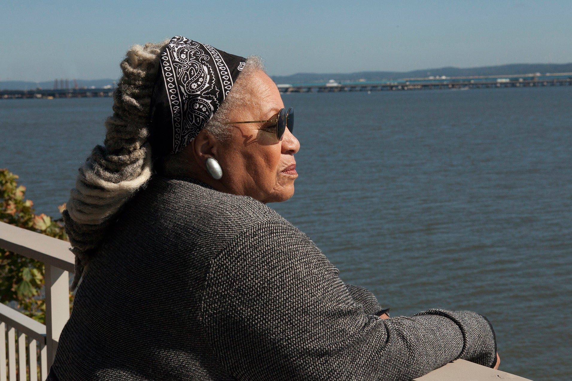 Toni Morrison, with greyed dreadlocks and sunglasses looks over a balcony across a large body of blue water. 