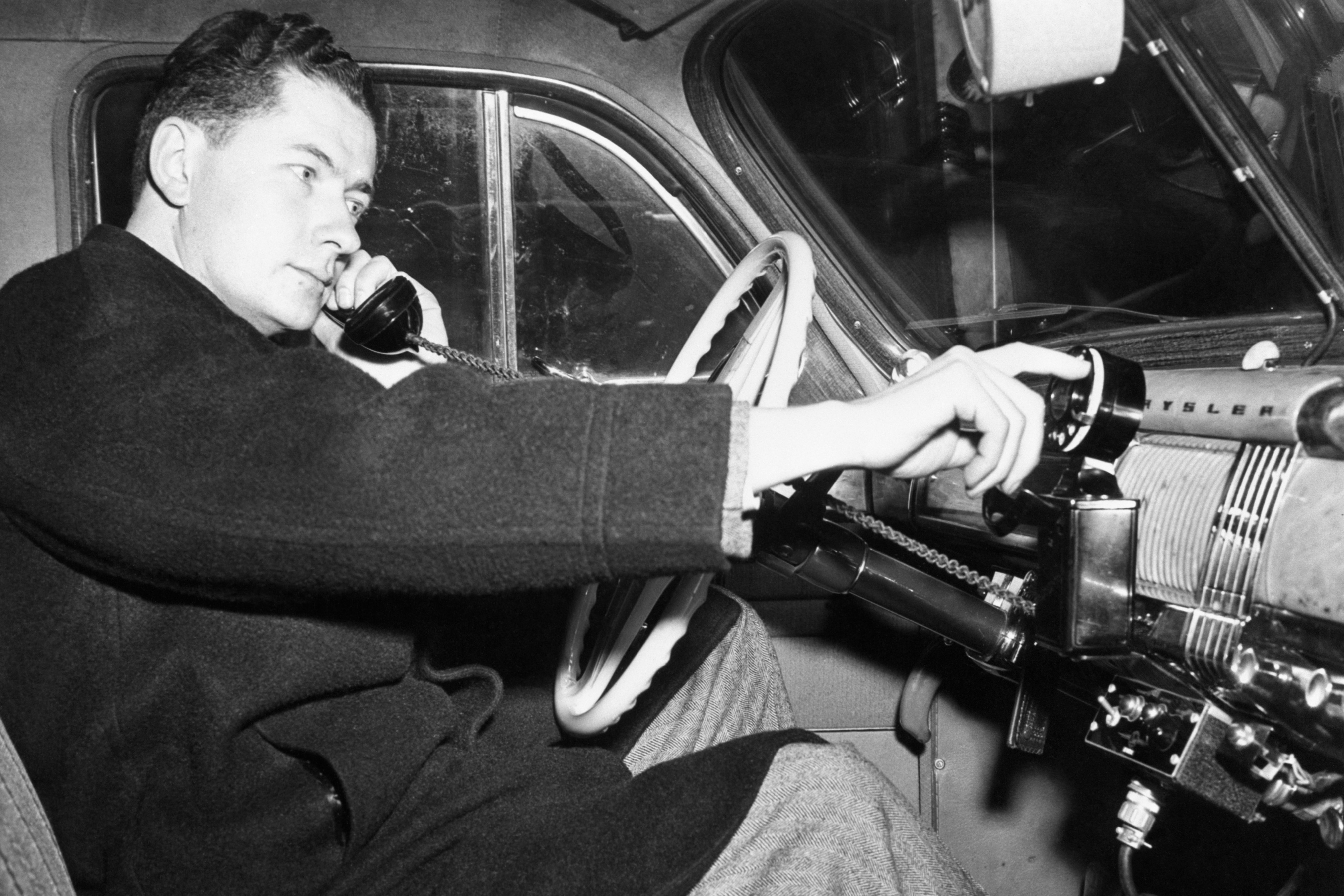 A man sits behind the wheel of a car with a phone to his ear as his finger uses a rotary dial.