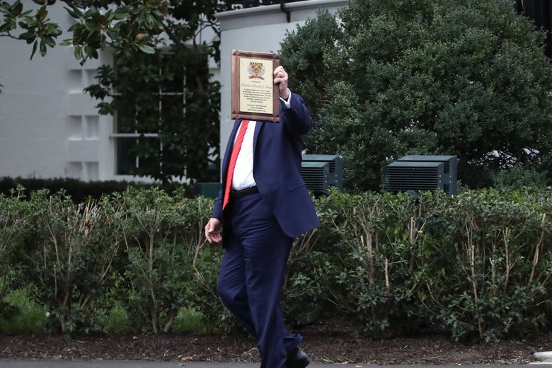 Trump, walking outside the White House, holds a plaque in the air such that it blocks his face.