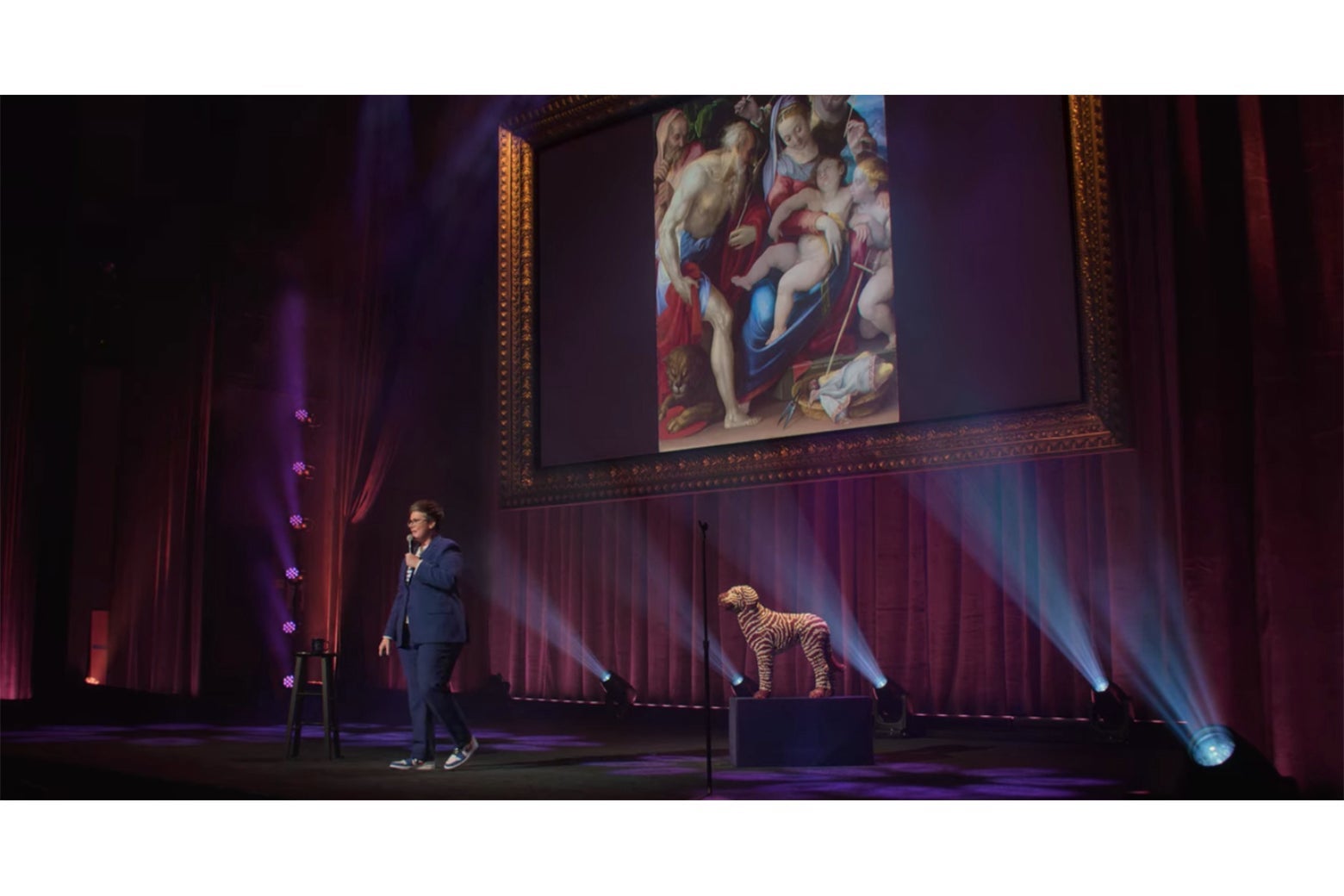 Hannah Gadsby onstage in front of a painting of the Holy Family in which the baby Jesus is gigantic for some reason.