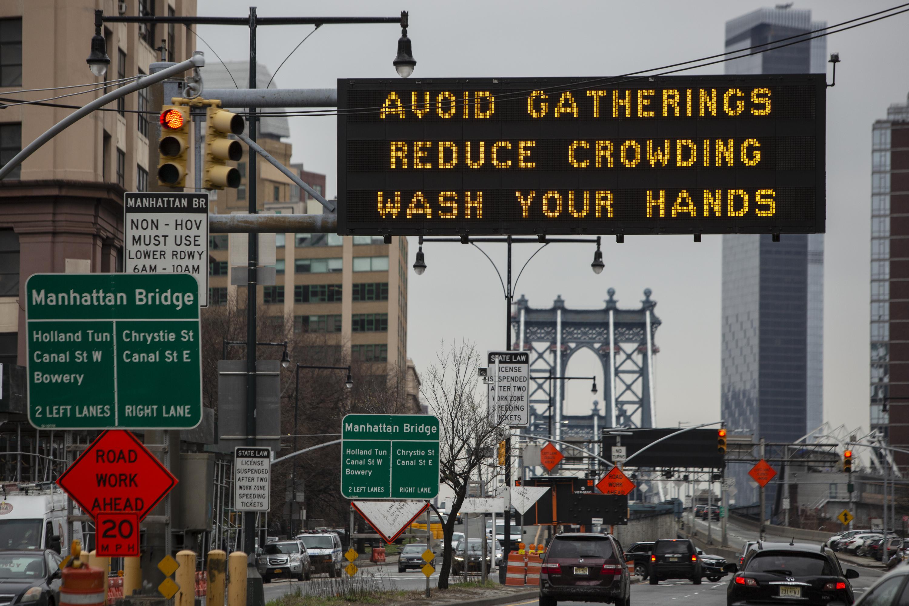 A street in New York, with signs, cars, and an announcement stating, "Avoid Gatherings. Reduce Crowding. Wash Your Hands." The Manhattan Bridge can be seen farther ahead.