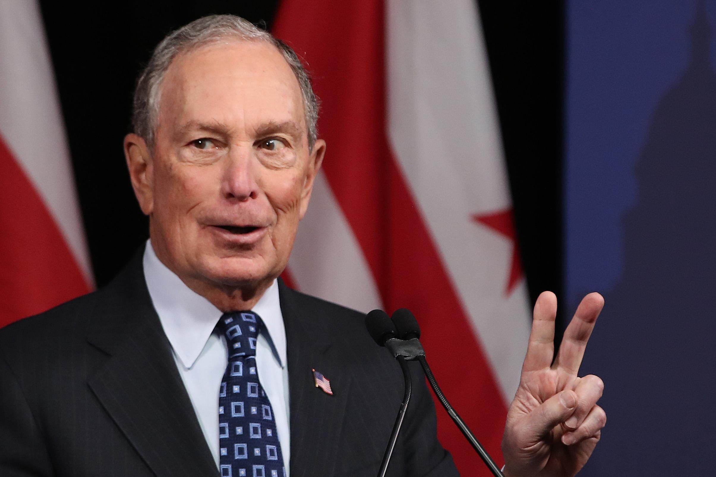 Michael Bloomberg stands in front of a microphone and flashes a peace sign.