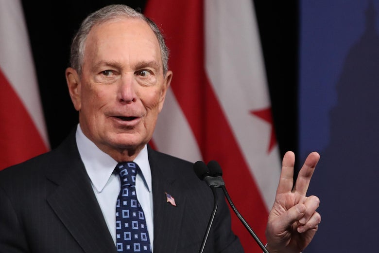 Michael Bloomberg stands in front of a microphone and flashes a peace sign.