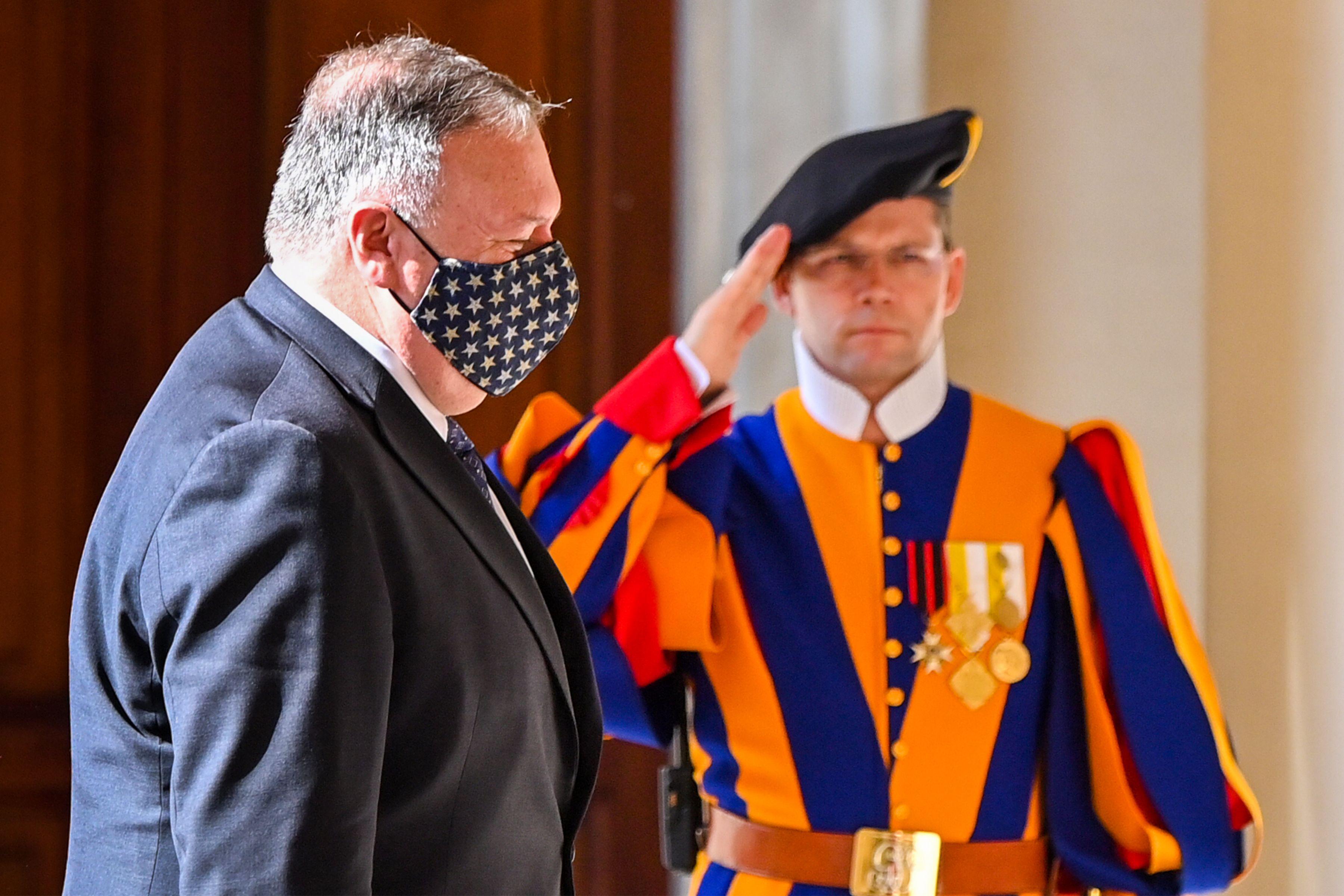 Mike Pompeo, wearing an American flag mask, walks in front of a Swiss Guard raising his arm in salute