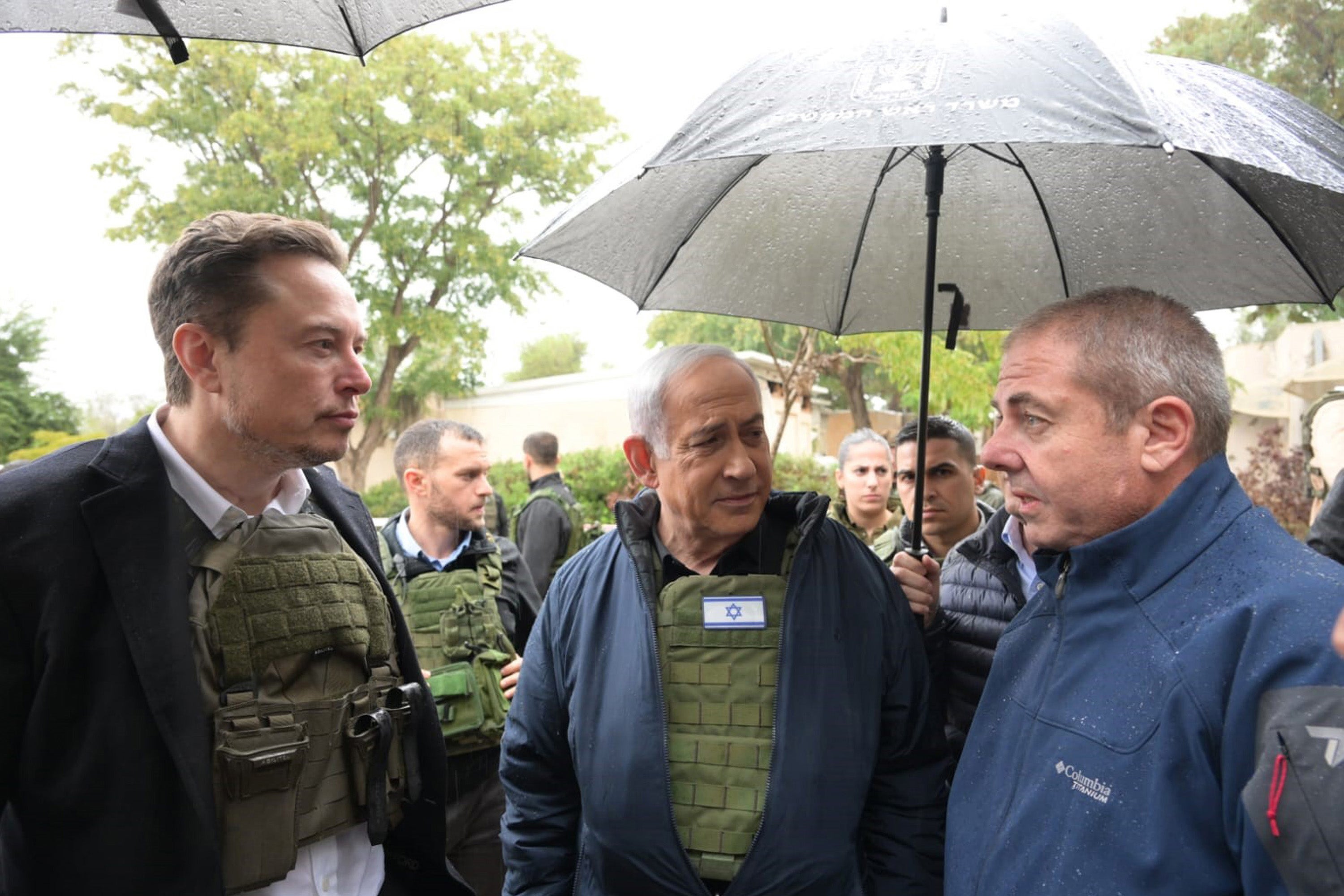 Musk and Netanyahu in bulletproof vests listening to an older man speak, under an umbrella.