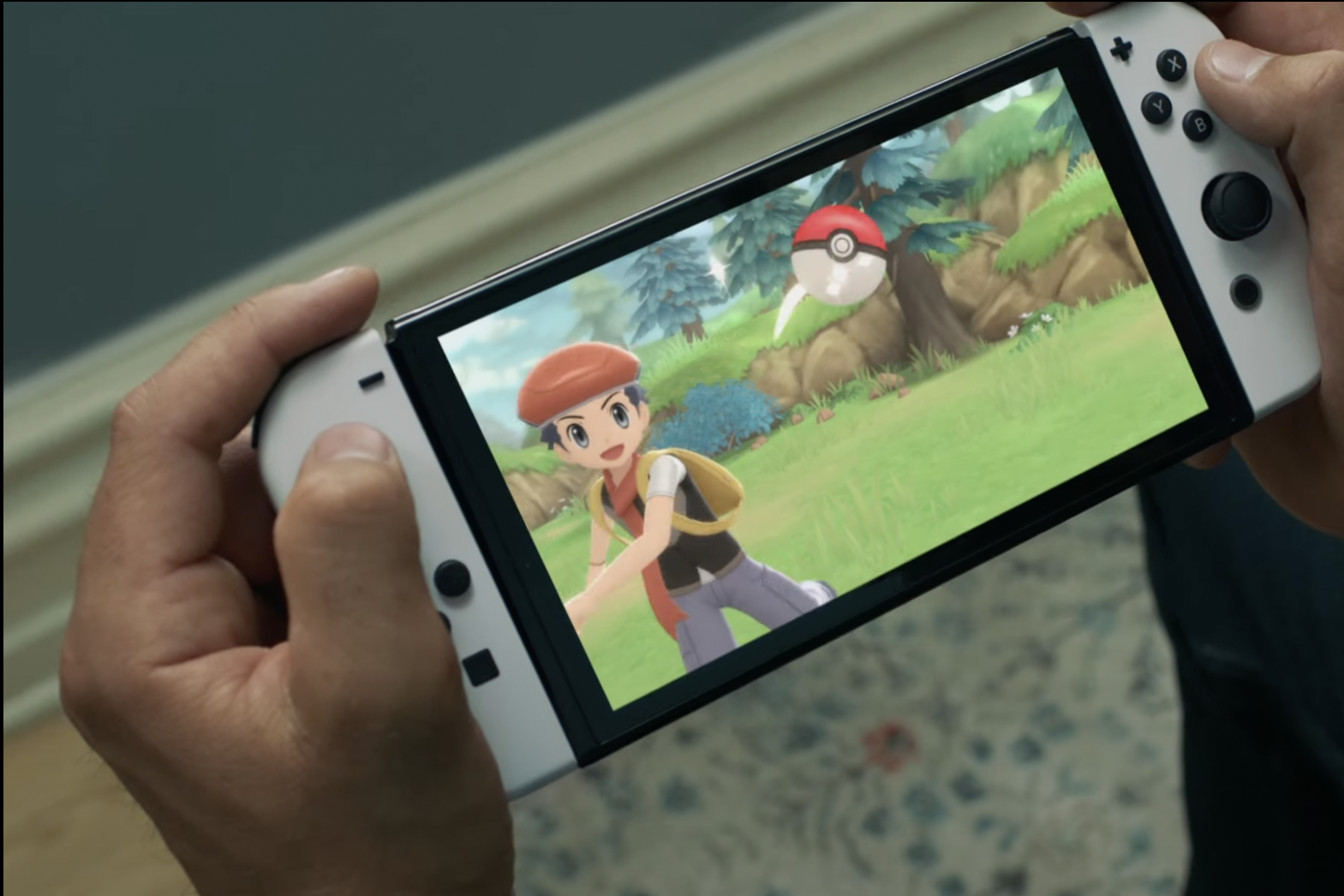 A pair of hands hold a Nintendo Switch console. On the screen is a boy wearing a red beret throwing a red-and-white ball across green grass. 