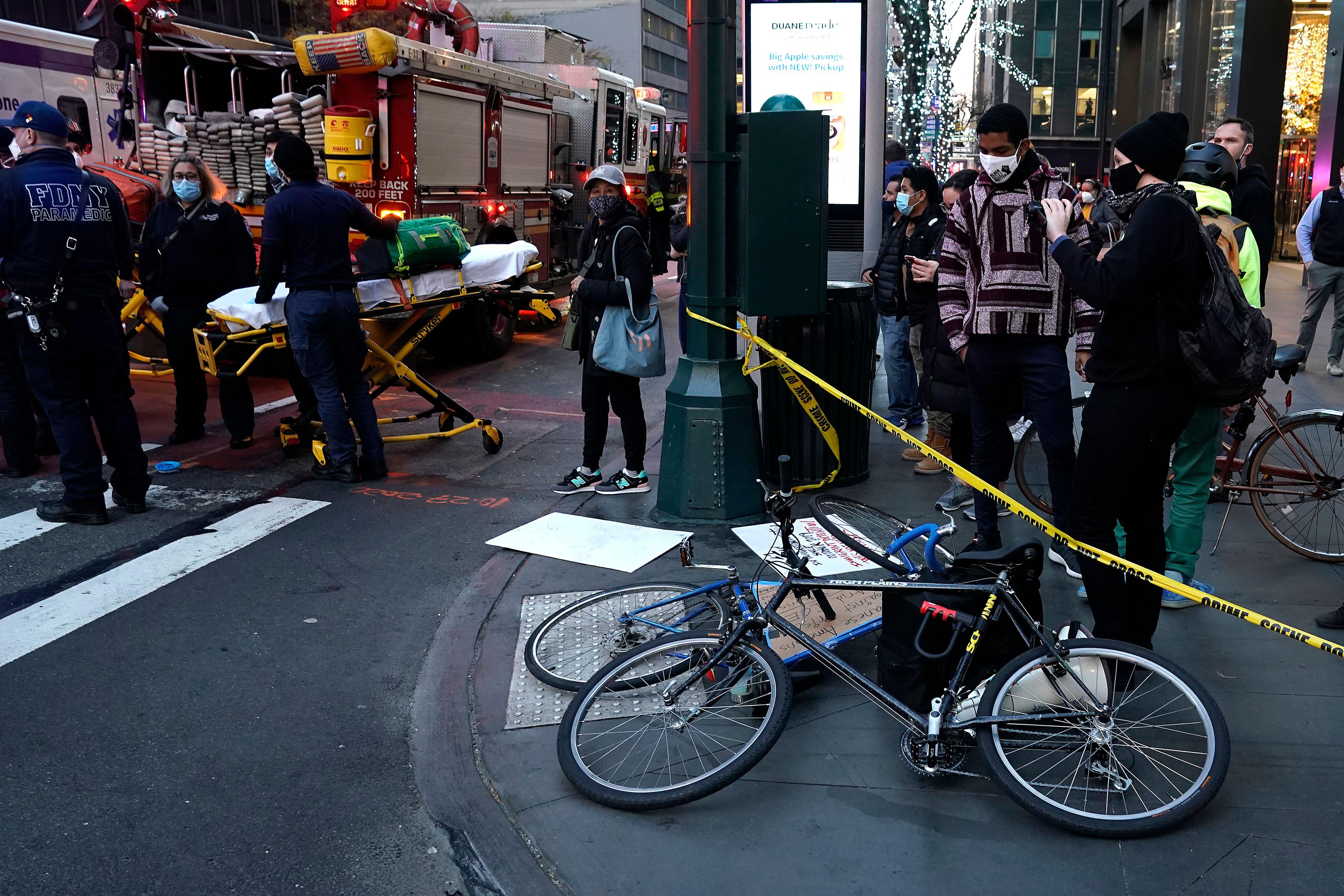 Two bikes are seen lying flat near police ticker tape. To the left, masked people hold a stretcher.