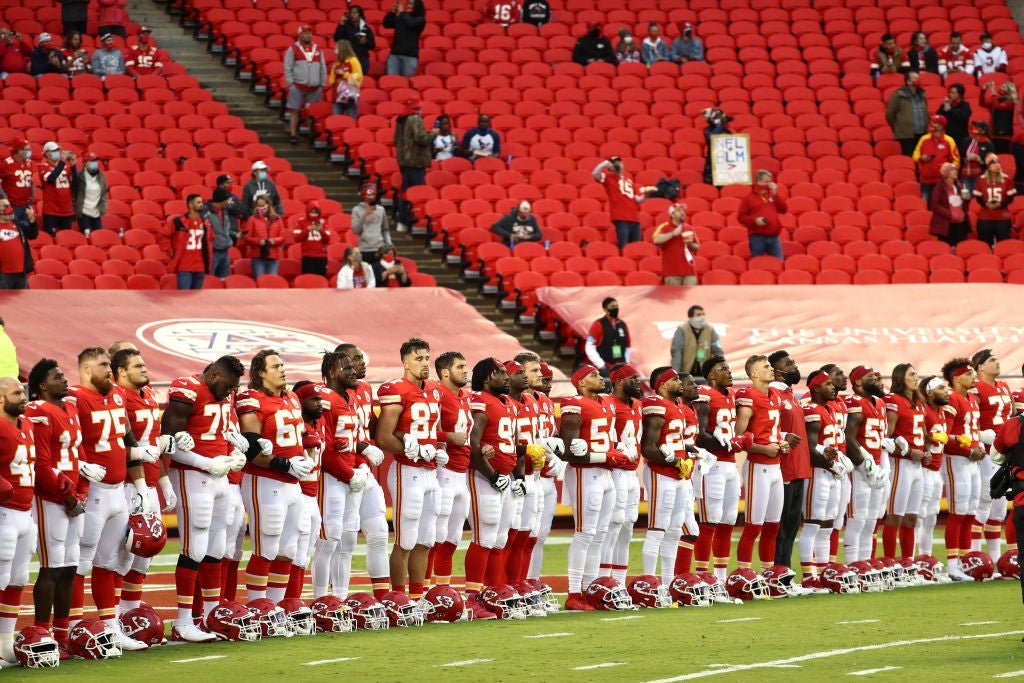 A line of players dressed in the Chiefs' red uniforms stands with locked arms in front of a sparse, socially distanced crowd.