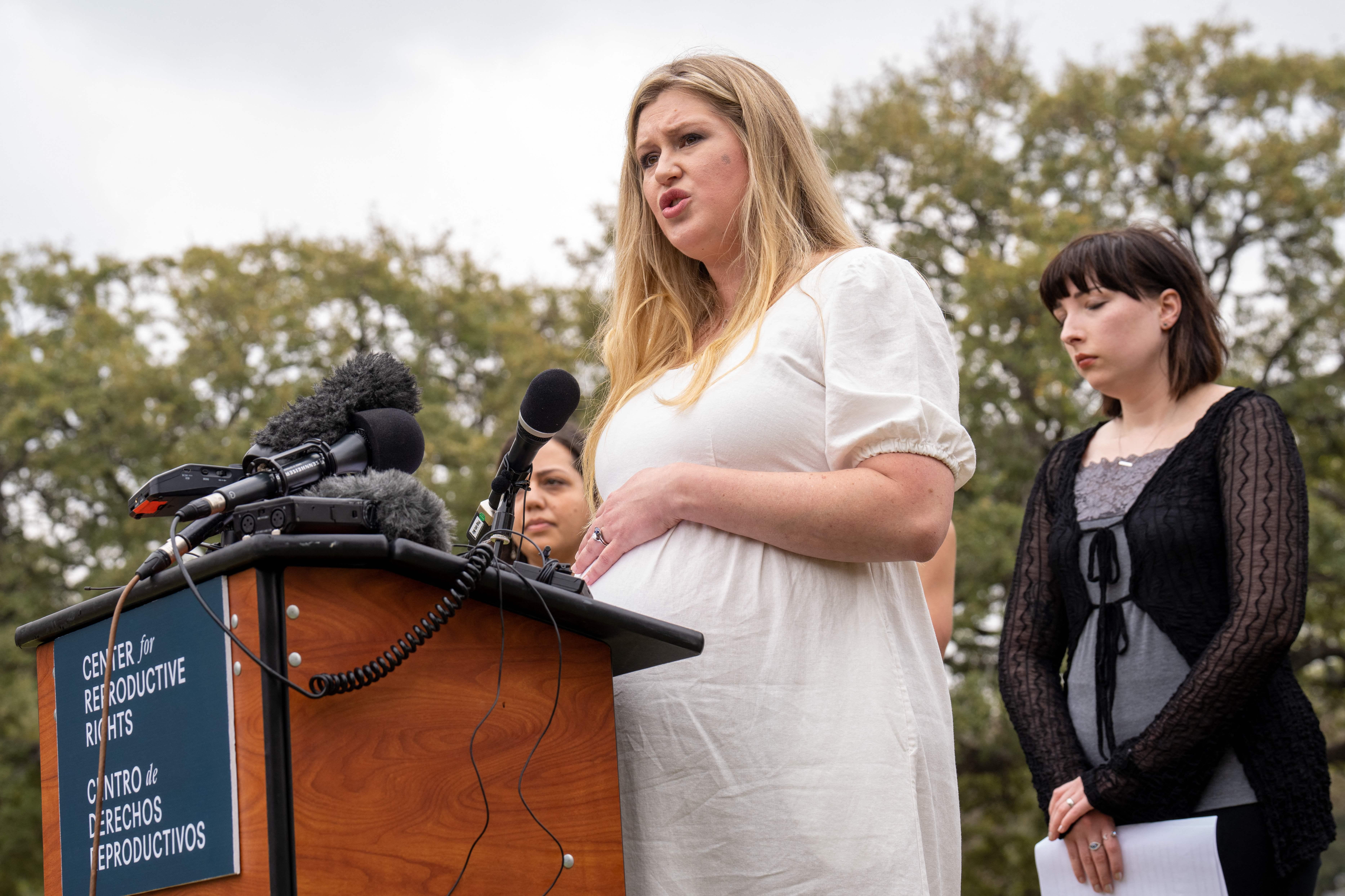 A visibly pregnant woman in all white speaks at a podium as another plaintiff stands behind her and looks down, appearing resigned, while holding a piece of paper.