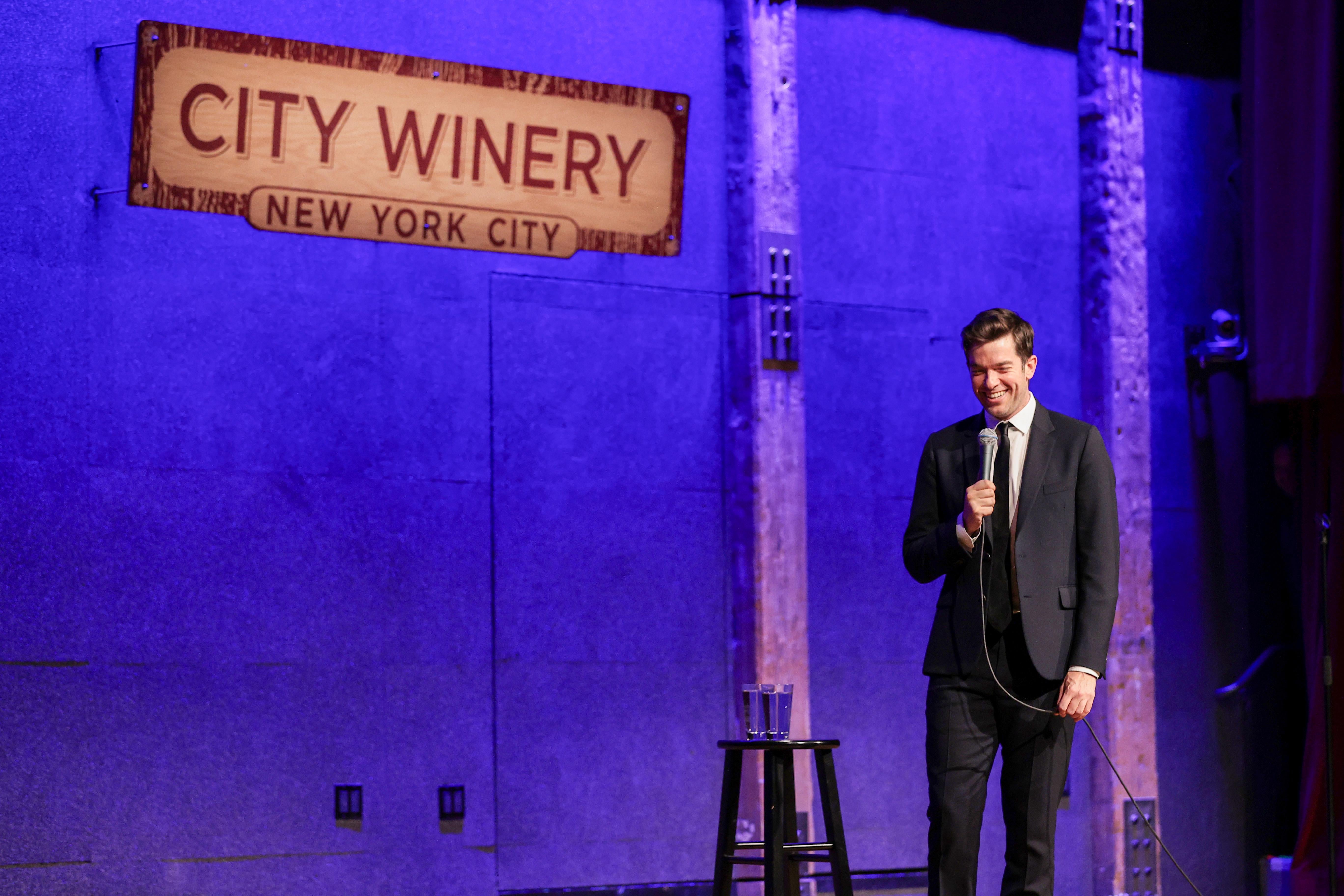 A man in a black suit and tie stands next to a stool with a cup of water on top of it. He's on a stage with a purple light behind him and a sign that says CITY WINERY above him. 