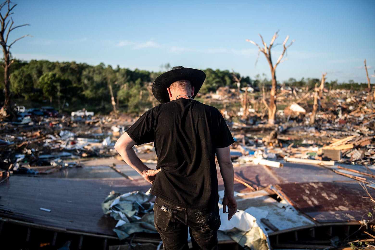 A white man wearing a brown cowboy hat stares down at the ground, with a pile of debris in the background.