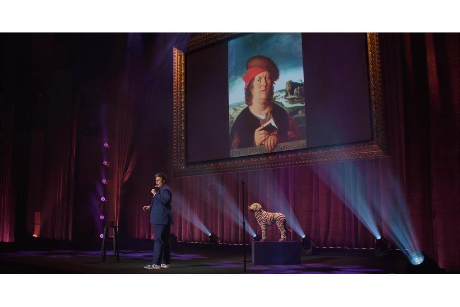 Hannah Gadsby stands in front of a projected image of a portrait of a not-very-bright-looking man dressed in red.