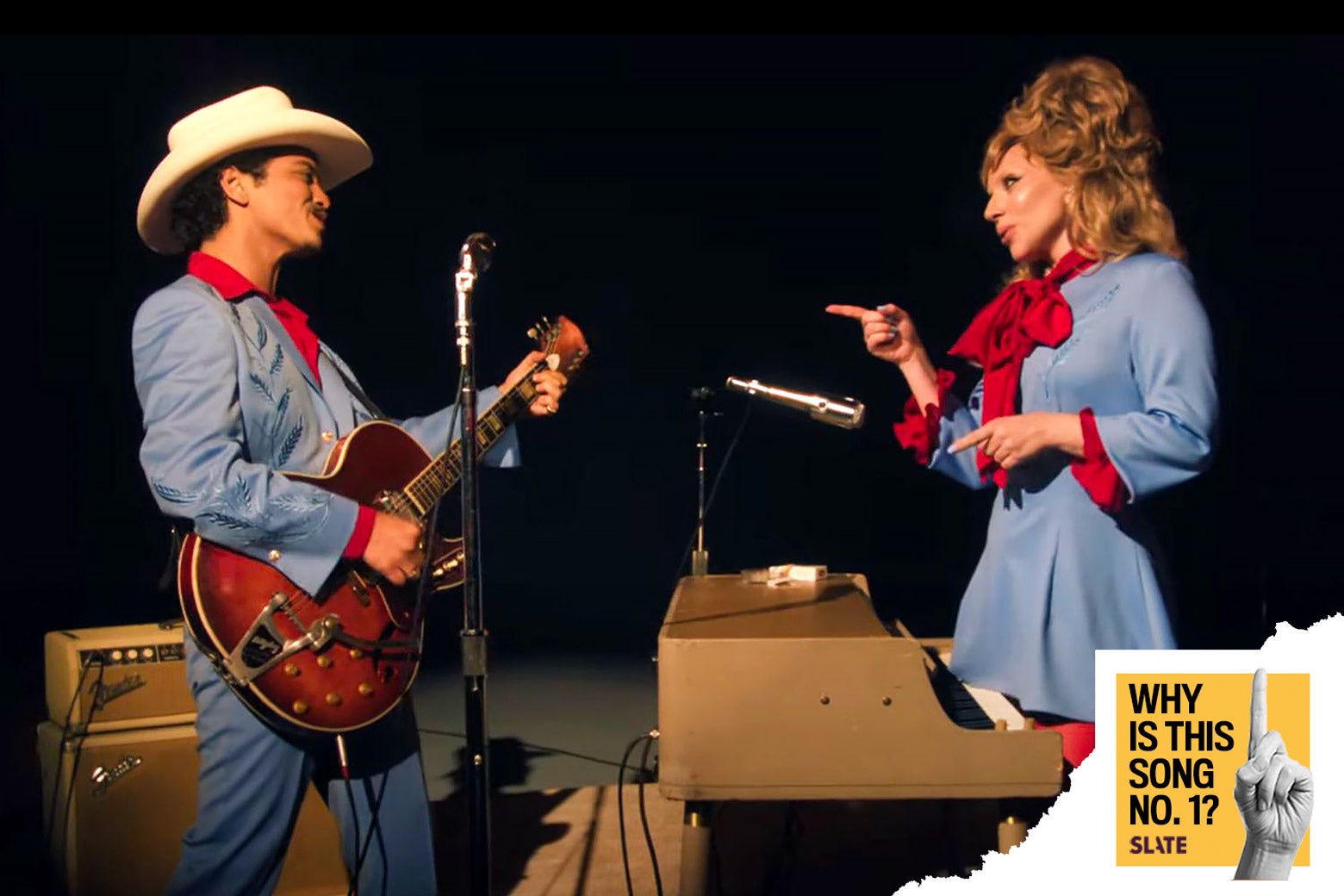 Mars and Gaga wear vintage baby blue and firetruck red country outfits. He looks lovingly at her while holding a vintage guitar in front of a tan amplifier. She points lovingly back at him while standing behind a vintage tan synthesizer.