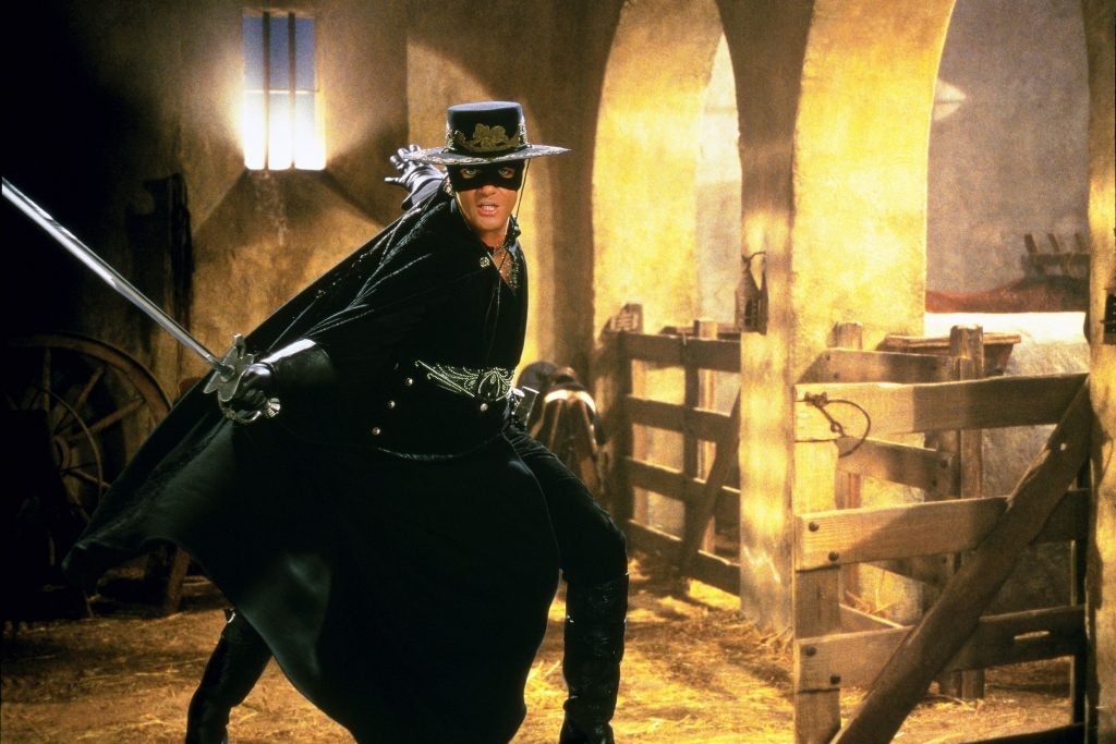 Antonio Banderas is standing mid-lunge in the classic Zorro black mask, hat, and cape while holding a sword out with an extended hand.