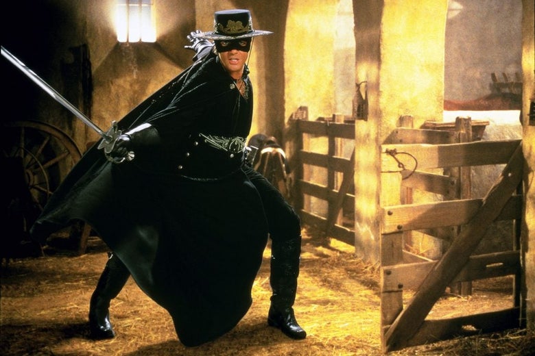 Antonio Banderas is standing mid-lunge in the classic Zorro black mask, hat, and cape while holding a sword out with an extended hand.