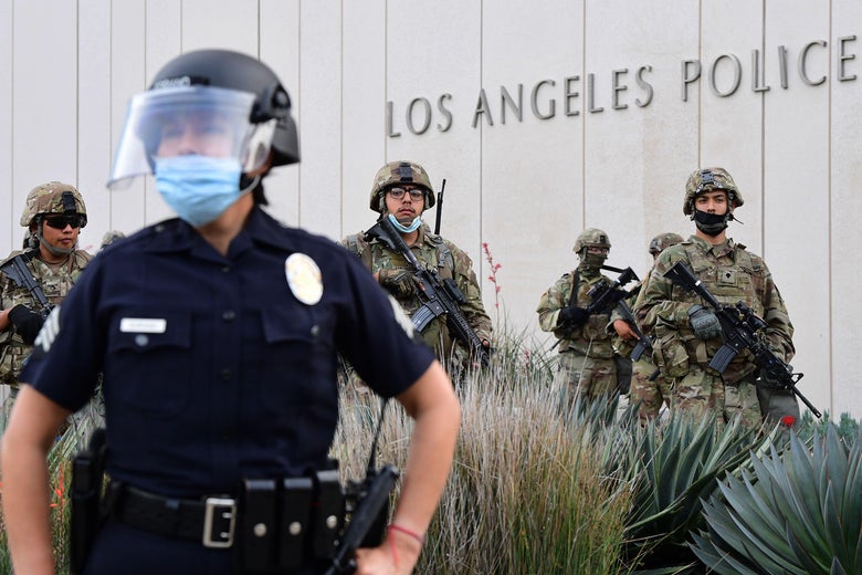 Un officier de police avec un écran facial tient des armes contre le côté devant un bâtiment avec un signe de membres de la police et de la garde nationale de Los Angeles en tenue camouflage et militaire.  