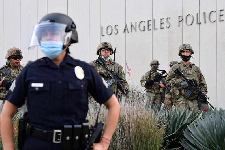 Un officier de police avec un écran facial tient des fusils contre le côté devant un bâtiment avec un signe de membres de la police et de la garde nationale de Los Angeles en tenue de camouflage et d'équipement militaire.