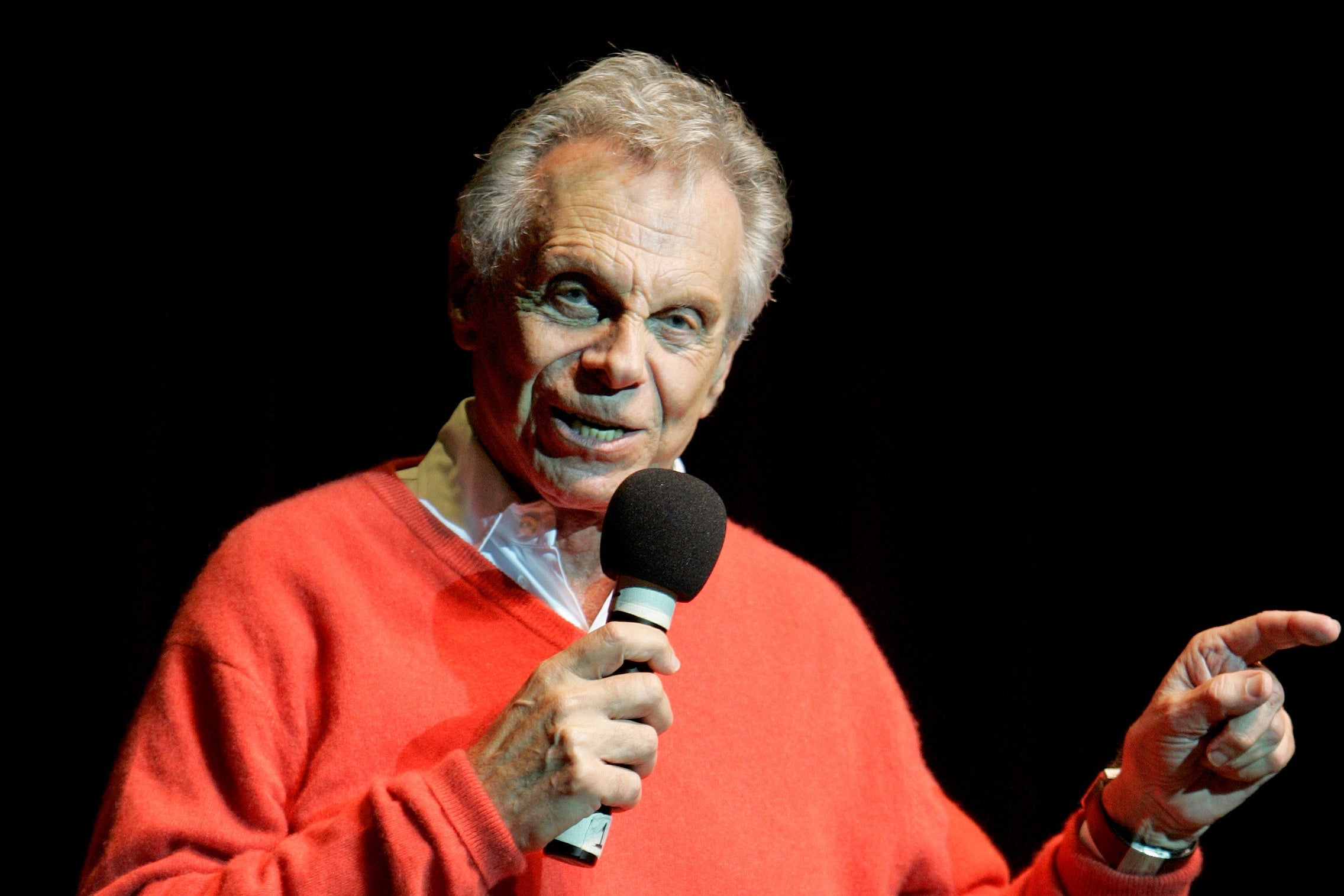 An older Mort Sahl in a red sweater onstage holding a microphone and pointing.