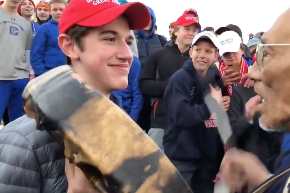 A teenage boy wearing a red MAGA hat smirks at Native American elder Nathan Phillips. A group of teenage boys cheer and join in the ridicule in the background.