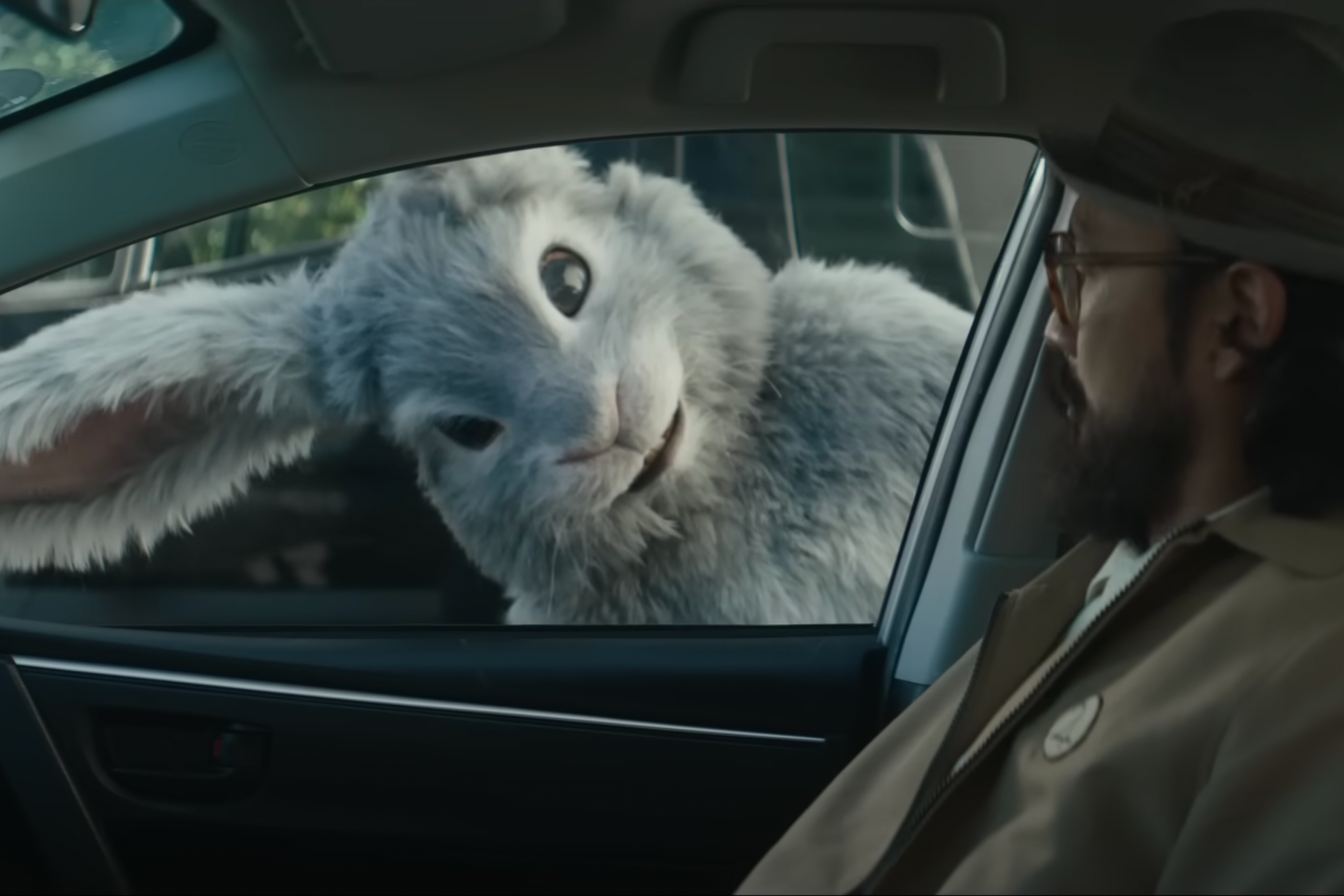 A human-sized rabbit peers into a car. The man inside appears confused.