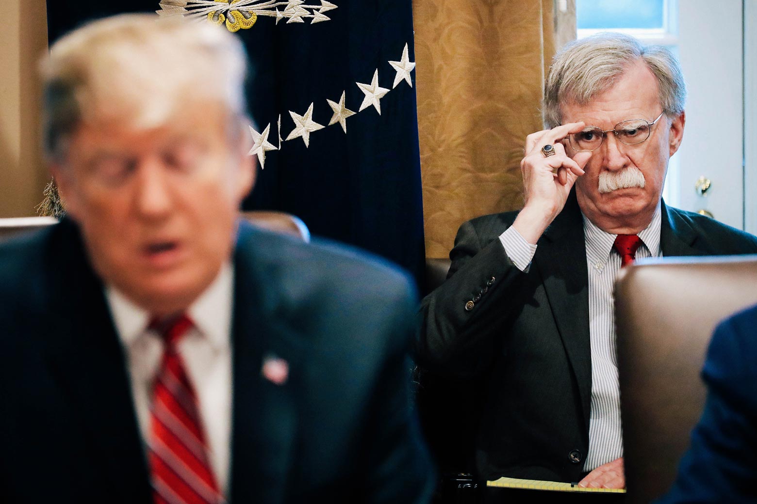 John Bolton, sitting in off to the side and behind Donald Trump, adjusts his glasses as Trump speaks.