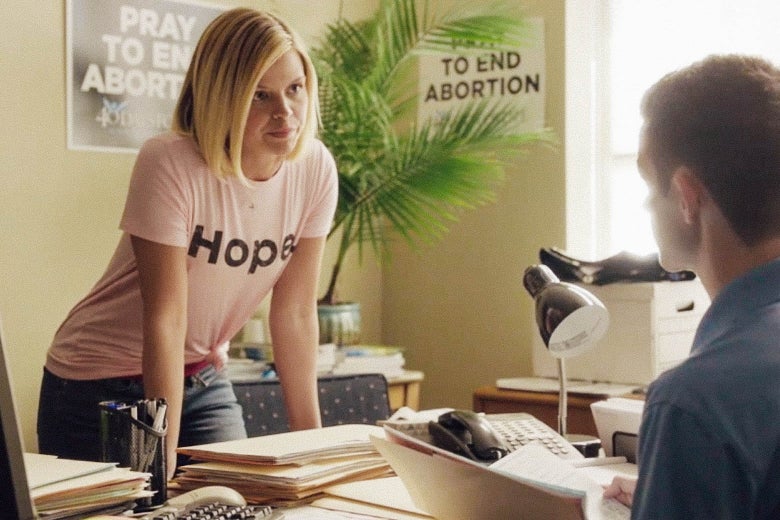 In this still from Unplanned, actress Emma Elle Roberts leans over an office desk while wearing a shirt that says "Hope." Jared Lotz's back is to the camera, as he is seated behind the desk.