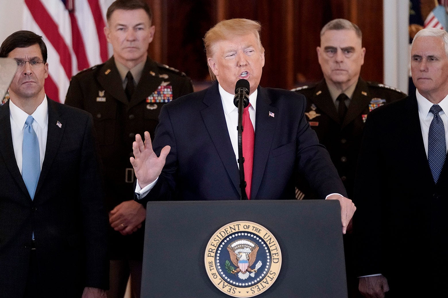 Donald Trump stands at a White House lectern, speaking and raising his hand, as the Joint Chiefs of Staff stand in the background.
