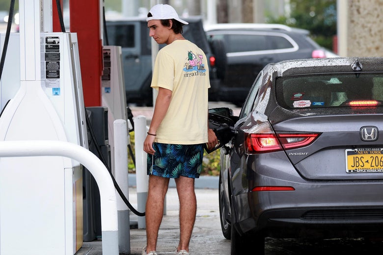 A young man in a T-shirt, a backward baseball cap, and shorts frowns at the pump as he fuels up his car at a gas station in Miami