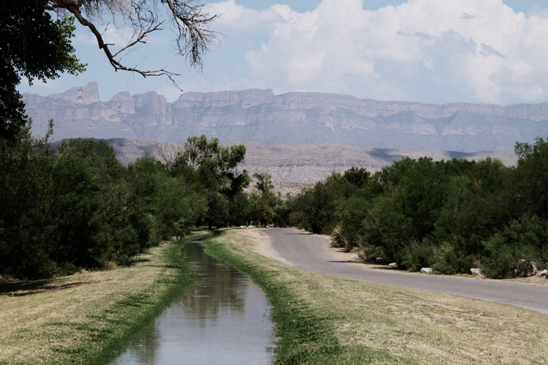 A canal with green banks runs along a road, with mountains in the distance.