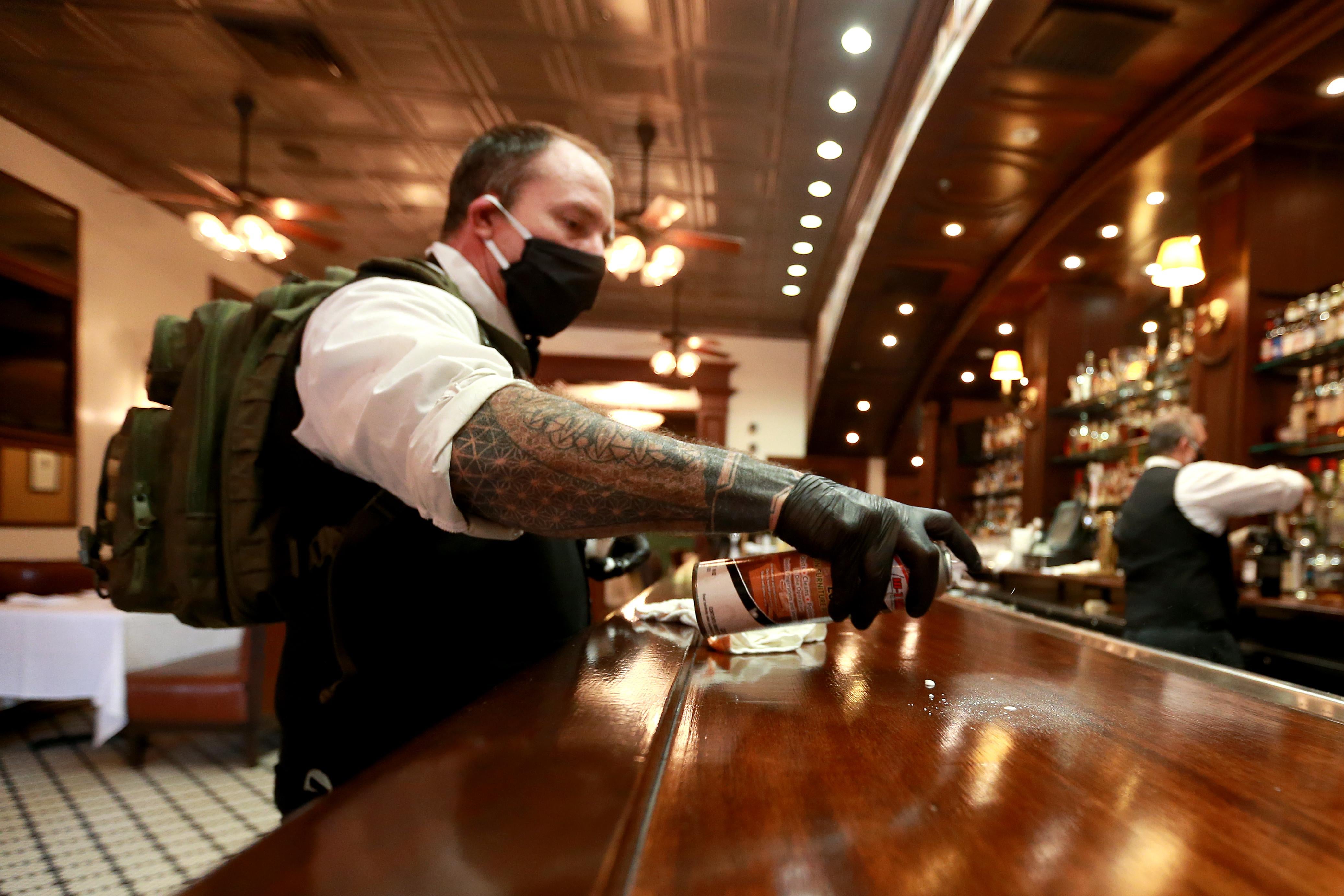 A bartender wearing a black mask and a backpack cleans the bar with disinfectant spray, while a bartender in the back mixes a drink.
